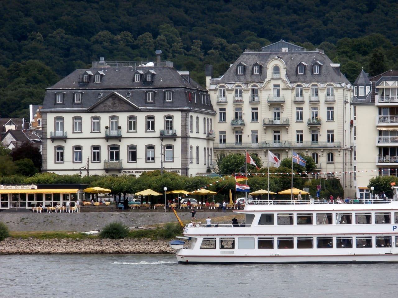 Das ehemalige Hotel Düsseldorfer Hof und sein Nachbarhaus an der baumgesäumten Rheinallee in Königswinter, davor ein Ausflugsschiff auf dem Rhein